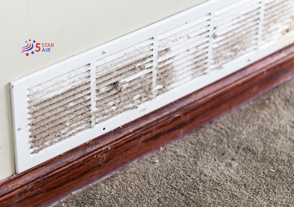 Close-up of a dirty HVAC return air vent covered in thick dust and debris on a white wall above wooden baseboards.