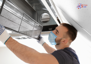 An HVAC technician wearing a face mask and protective gloves inspects a large, dusty air filter from a ceiling-mounted ventilation unit